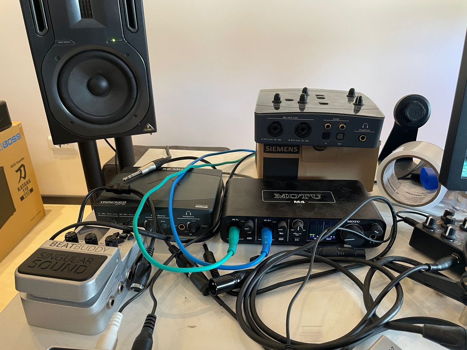 A cluttered desk holding an E-MU Tracker Pre, a MOTU M4 audio interface, a studio monitor, a BeatBuddy pedal and a tangle of patch cables
