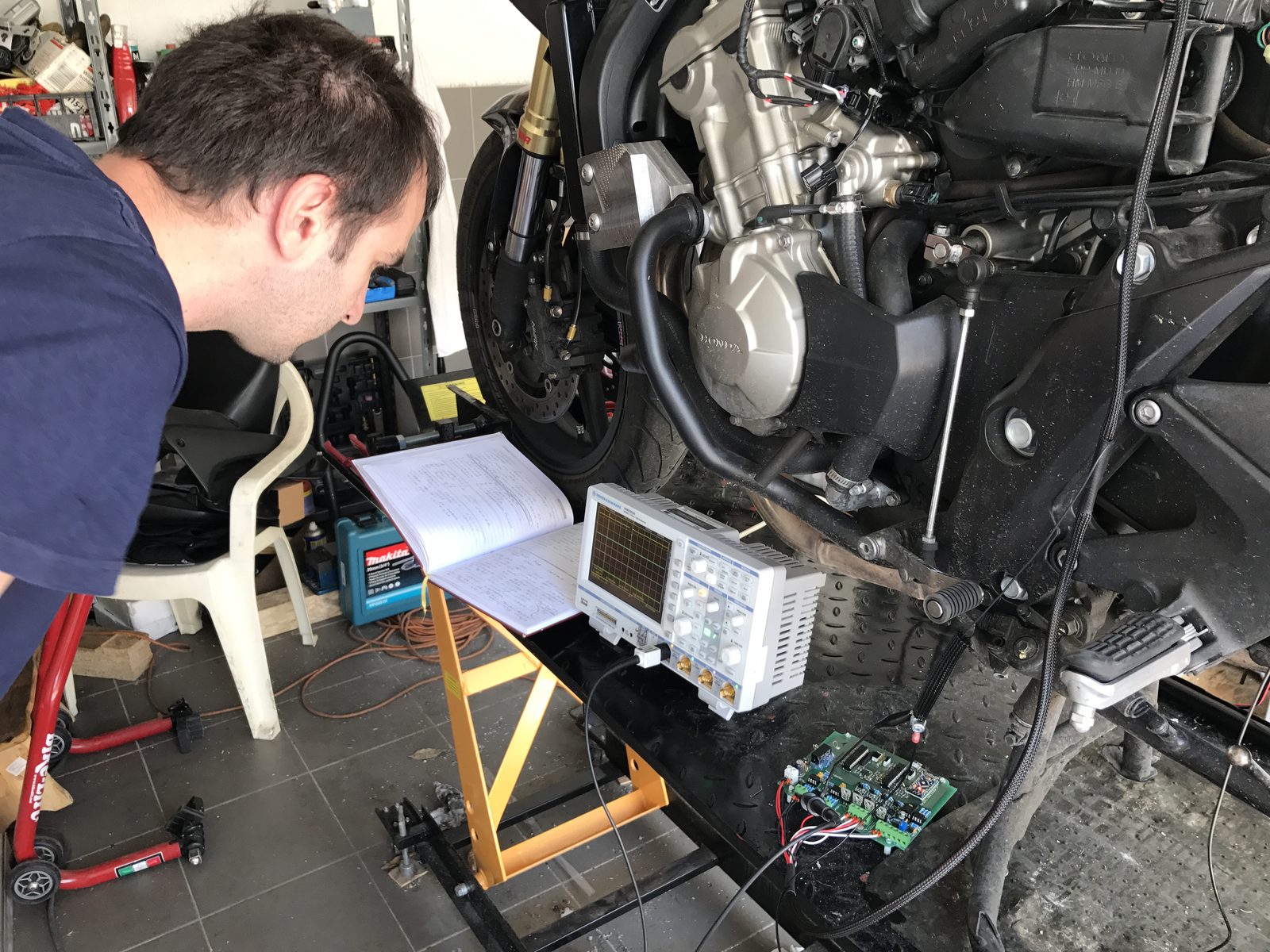 Oscilloscope connected to a motorcycle on a workshop stand with the engine running, notebook of handwritten measurements alongside
